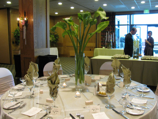 Ein Tisch mit einer weißen Decke, Tellern, Besteck, einem Glas und Stühlen drumherum, einer grünen Wand dahinter, einem Mann in einem schwarzen Blazer, einer stehenden Frau und einer Tür mit der Aufschrift 'Ausgang' sowie zwei Männern dahinter.