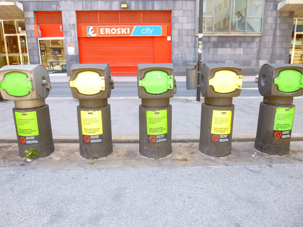 Eine Reihe von Parkautomaten entlang einer Straße mit einem Gebäude, das Glasfenster und eine Rolllade mit Text im Hintergrund sowie einen nahen Müllcontainer.