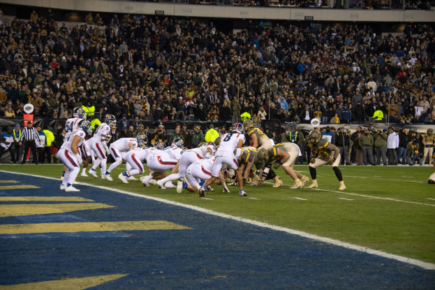 Ein Fussballspiel in einem Stadion mit Spielern in Sportkleidung und Helmen auf dem Feld, Zuschauern, Gel├Ąndern und Tafeln mit Text im Hintergrund.