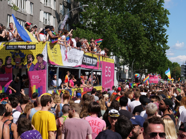 Eine große Menge marschiert während des Berlin Pride 2019 eine Straße entlang, viele tragen Mützen und Schutzbrillen, einige halten Fahnen, mit Bannern, Laternenmasten, Bäumen und Gebäuden im Hintergrund bei einem bewölkten Himmel.