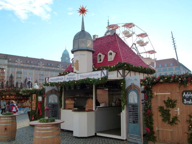 Kleines rotes Dachgebäude mit einem Riesenrad davor, umgeben von Festbesuchern, Fässern, Pflanzen, Schildern und Dekorationen, vor einer Kulisse aus Gebäuden, Bäumen und einem bewölkten Himmel während des Oktoberfestes in München, Deutschland.