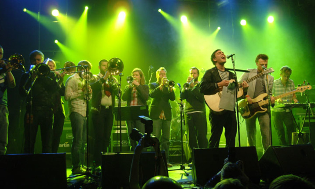 Eine Gruppe von Musikern performs auf der Bühne mit Mikrofonen, Scheinwerfern und einer Kamera, während das Publikum zuschaut.