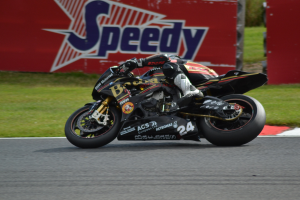 Eine Person in Helm und Schutzkleidung fährt ein Motorrad auf einer Rennstrecke, im Hintergrund ist Gras und eine Texttafel zu sehen.