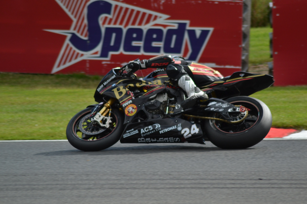 Eine Person in Helm und Schutzkleidung fährt ein Motorrad auf einer Rennstrecke, im Hintergrund ist Gras und eine Texttafel zu sehen.