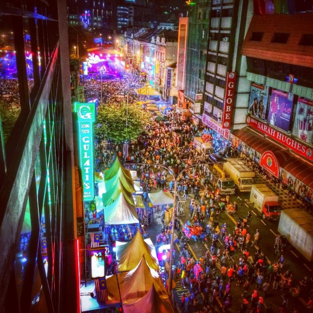 Ein lebhaftes Volksfest-Szenario mit einer großen Menschenmenge auf der Straße, umgeben von Zelten, Geschäften, Gebäuden und Bannern, mit einer hell erleuchteten Fläche im Hintergrund.