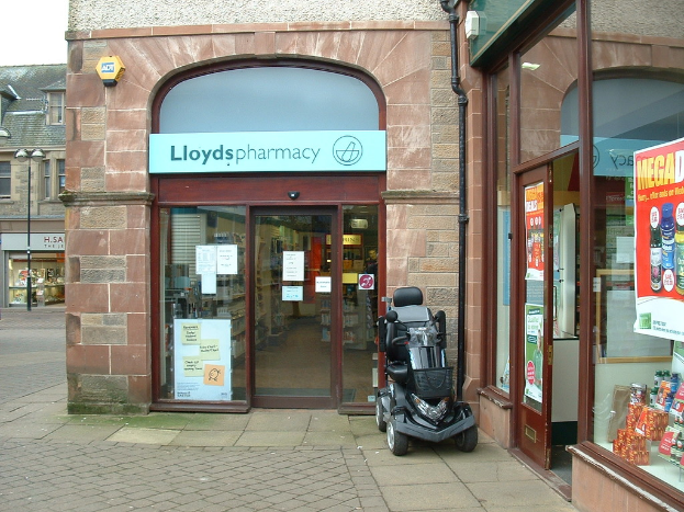 Eine Apotheke mit einem vor ihr geparkten Fahrzeug und einem Gebäude im linken Eck.