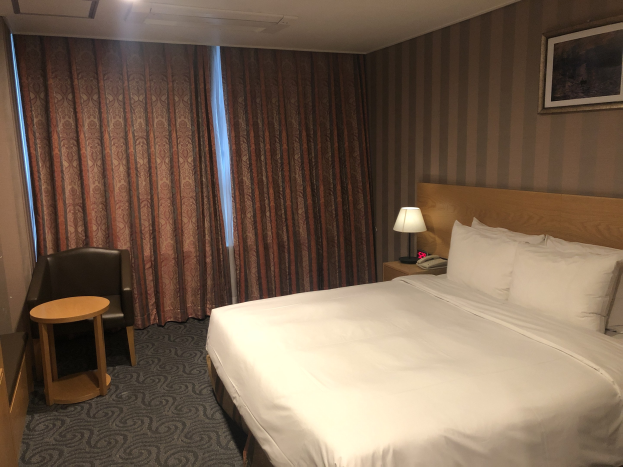 Hotelzimmer mit einem großen Bett, einem Stuhl, einem Tisch, einem Telefon, einer Lampe, einem Foto auf der Wand, einem Fenster mit Vorhängen und einer Decke mit Leuchten.