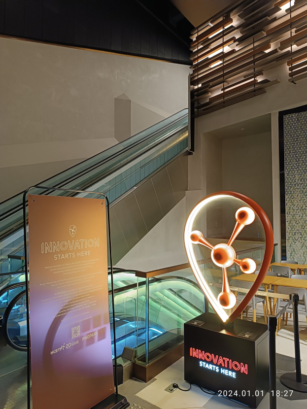 Großes Schild in der Mitte eines Gebäudes mit einer Rolltreppe im Hintergrund, Tische und Stühle auf der rechten Seite und eine Tafel auf der linken Seite mit der Aufschrift "Innovation Starts Here", die auf die HSTP20 Expo hinweist.