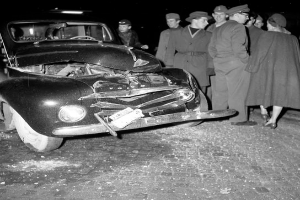 Eine Schwarz-Weiß-Szene eines beschädigten Autos am Straßenrand, mit einer Gruppe von Menschen, die es besichtigen.