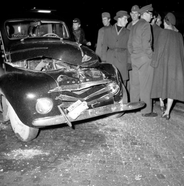 Eine Schwarz-Weiß-Szene eines beschädigten Autos am Straßenrand, mit einer Gruppe von Menschen, die es besichtigen.