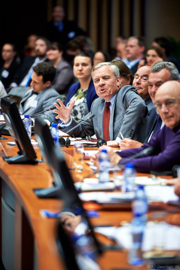 Eine Gruppe von Menschen sitzt um einen Tisch mit Bildschirmen, Flaschen, Gläsern, Papieren und Headsets, wobei die Person in der Mitte einen Anzug trägt und der Hintergrund unscharf ist.