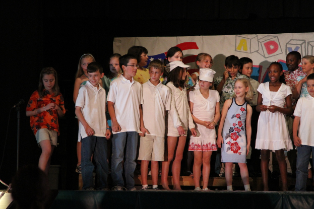 Mehrere Kinder auf einer Bühne, zwei in der Mitte mit Mützen, ein Mikrofonständer links und ein Banner im Hintergrund.