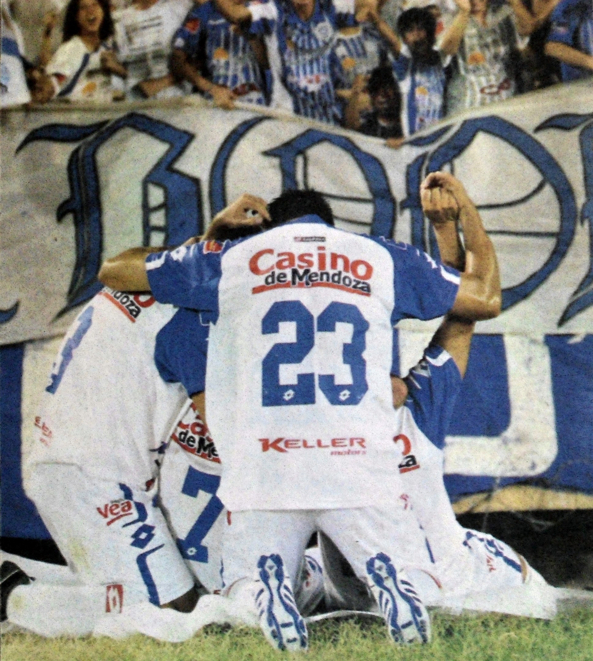 Eine Gruppe von Fußballspielern, die ein Tor feiern, vor jubelnden Zuschauern auf dem Boden kniend, mit einem teilweise sichtbaren Banner im Hintergrund.