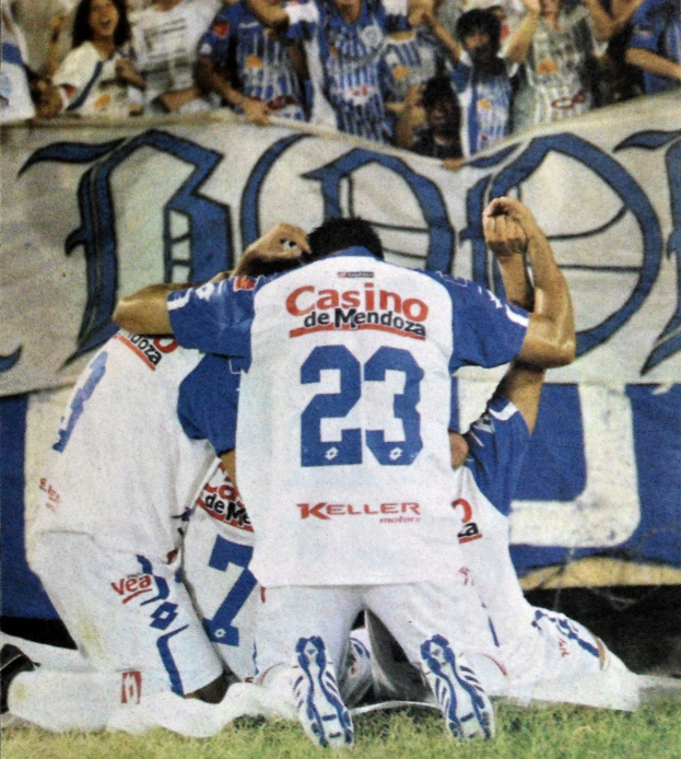 Eine Gruppe von Fußballspielern, die ein Tor feiern, vor jubelnden Zuschauern auf dem Boden kniend, mit einem teilweise sichtbaren Banner im Hintergrund.