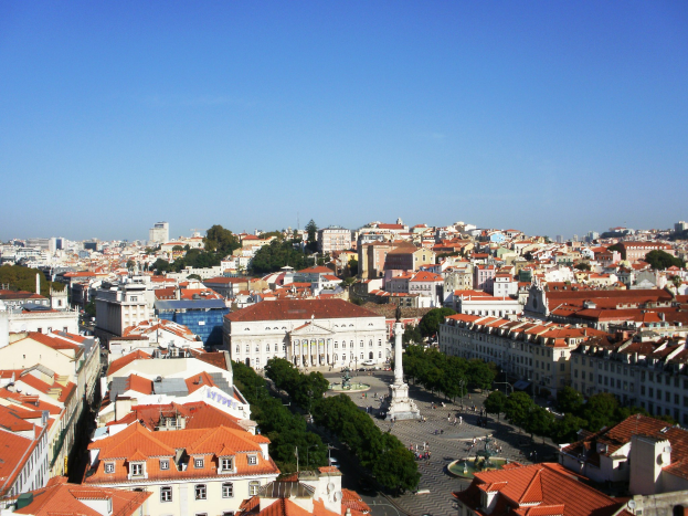 Blick auf Lissabon von einem Hügel aus, der Gebäude, Bäume, eine Statue auf einem Sockel, Menschen auf der Straße und den Himmel im Hintergrund zeigt.
