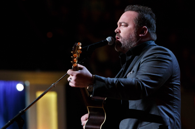 Ein Mann in einem Anzug spielt Gitarre und singt in ein Mikrofon auf einer schwach beleuchteten Bühne, mit Dierks Bentley im Hintergrund.