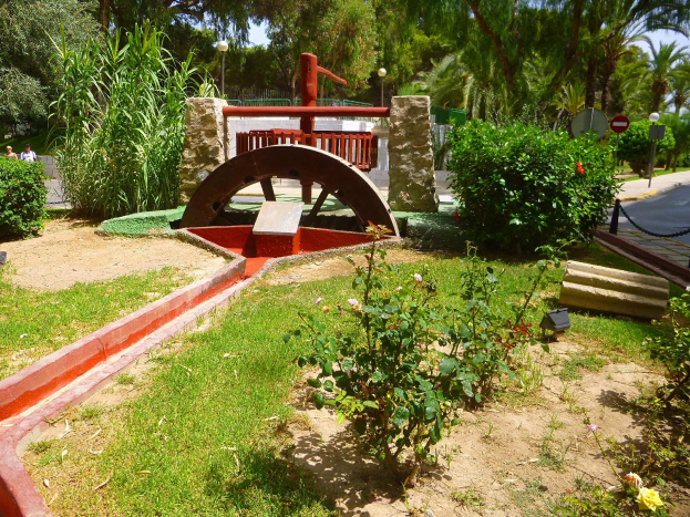 Ein Minigolfplatz in einem Park, umgeben von Grün, mit einer Straße, Fußgängern, Schildern und einem klaren blauen Himmel im Hintergrund.