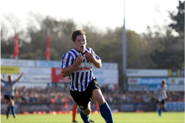 Ein Mann in einem blauen und weißen gestreiften Hemd rennt mit einem Fussball auf einem grünen Feld während eines Spiels von Huddersfield Town gegen Swansea City, mit Zuschauern, Tafeln, Fahnen und Bäumen im Hintergrund unter einem klaren blauen Himmel.