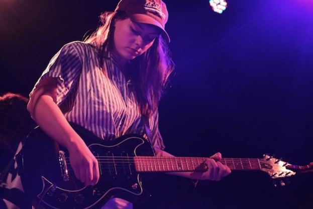 Eine Frau mit einer Mütze spielt Gitarre auf der Bühne eines Musikkonzerts, während zahlreiche Lichter den Hintergrund erhellen.