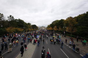 Eine große Gruppe von Menschen marschiert auf einer von Bäumen gesäumten Straße in Berlin, hält Kameras in der Hand, mit einem Gebäude und einem klaren Himmel im Hintergrund.