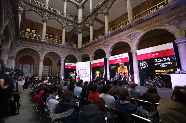 Eine Gruppe von Menschen, die auf einer Reihe von Stühlen vor einem Publikum sitzen, wobei einige Kameras halten, während einer Pressekonferenz für das CDMX 2017-Event, mit einem Podium mit einem Mikrofon in der Mitte, Bannern mit Text und Bildern, Säulen, Geländern, Fenstern und einer Wand im Hintergrund.