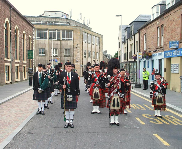 Eine Gruppe von Menschen, einige stehen auf der Straße und andere spielen Musikinstrumente, mit Gebäuden, Plakaten, Masten, Bäumen und Reifen im Hintergrund.