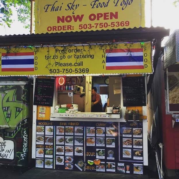 Eine Straßenansicht mit verschiedenen Schildern, Speisekarten, Werbetafeln und Getränkedosen sowie Gewürzen und Kondimenten in Plastikbehältern draußen.