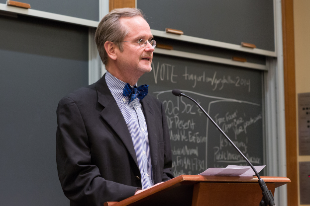 Ein Mann in einem schwarzen Anzug steht an einem Rednerpult und spricht in ein Mikrofon, mit einer Tafel im Hintergrund, auf der Text zu sehen ist.