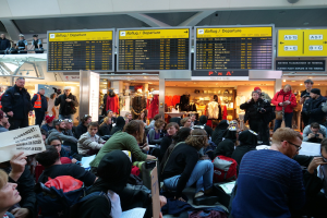 Eine große Gruppe von Menschen an einem Flughafen, einige sitzen mit Taschen und Papieren, andere stehen, mit Texttafeln, Schaufensterpuppen in Kleidern und Deckenleuchten im Hintergrund, was auf eine Protestveranstaltung hinweist.