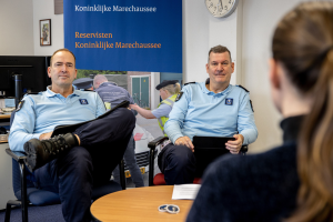 Zwei Polizeibeamte sitzen an einem Tisch mit Papieren und Gegenständen und sprechen mit einer Frau, im Hintergrund eine Wand mit einem Banner, einer Uhr und Fotorahmen, sowie ein weiterer Tisch mit einem Monitor, Tastatur und Maus links daneben.