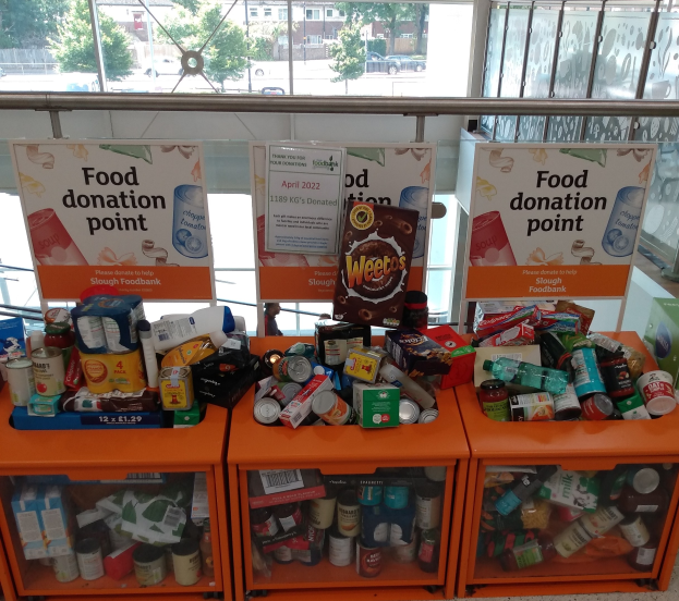 Orange Schübe mit Lebensmittelspenden, darunter Flaschen, Dosen und Kisten, mit Informationsschildern, durch Glasfenster mit Fahrzeugen, Bäumen, Gebäuden und Himmel.