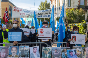 Eine Gruppe von Menschen mit Masken und Schildern und Spruchbändern steht vor einem Zaun mit Bildern und Text, protestiert in Istanbul, Türkei, mit Gebäuden, Bäumen und einem Laternenpfahl im Hintergrund.