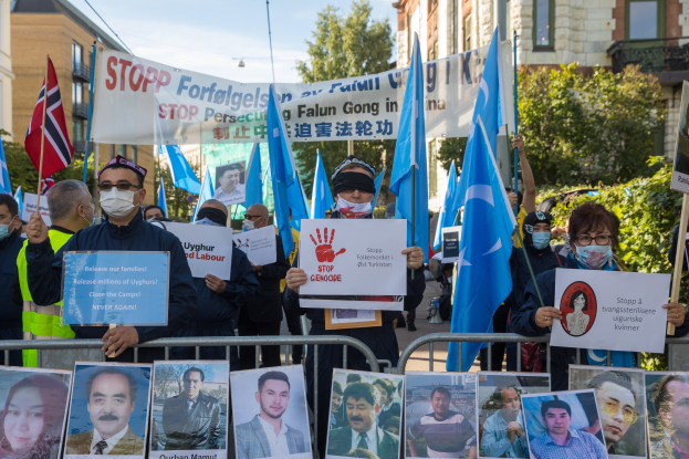 Eine Gruppe von Menschen mit Masken und Schildern und Spruchbändern steht vor einem Zaun mit Bildern und Text, protestiert in Istanbul, Türkei, mit Gebäuden, Bäumen und einem Laternenpfahl im Hintergrund.