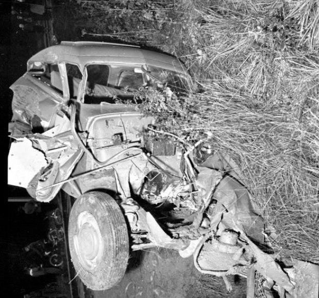 Ein Schwarz-Weiß-Foto eines schwerbeschädigten Autos am Straßenrand mit einer zerbeulten Front, zersplitterten Fenstern, einem Baum im Hintergrund und verstreuten Gegenständen rund um die Unfallstelle.