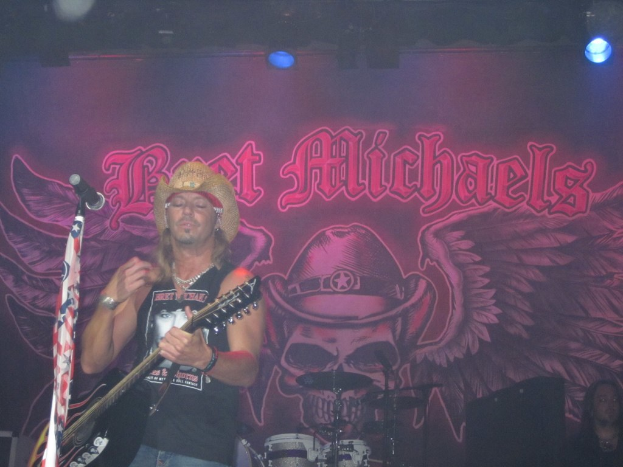 Eine musikalische Konzertsituation mit einem Gitarristen links, der in der Nähe eines Mikrofons spielt, einer Person, die in der unteren rechten Ecke sitzt, einem Banner im Hintergrund und Drogen, die in der Mitte unten platziert sind, mit Beleuchtung von oben.