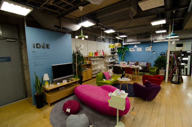 Eine Ladeneinrichtung mit Haushaltsgeräten und Möbeln, darunter ein Fernseher auf einem Tisch, eine rosafarbene Couch, ein Sofa, zwei Stühle um einen kleinen Hocker, ein Esstisch mit Stühlen, ein großes Sofa im Hintergrund und verschiedene Gegenstände auf Regalen, mit Deckenbeleuchtung.