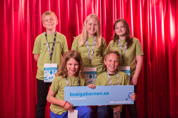 Eine Gruppe von Kindern mit ID-Karten, wobei ein Junge und ein Mädchen ein Schild mit Text halten und Vorhänge oben auf dem Bild zu sehen sind.