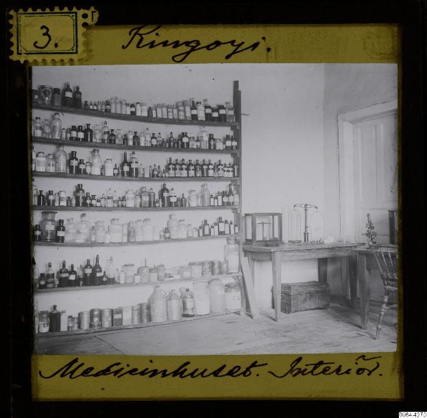 Schwarz-weiß-Foto eines alten Apothekeninneren mit Regalen voller Flaschen, einem Tisch mit Gegenständen, einem Stuhl, einer Tür und einer Wand mit Text unten.