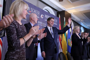 Eine Gruppe von Menschen, die vor Fahnen stehen und jubeln, mit einem Tisch voller Gegenstände rechts daneben und Stühlen, einer Fahne und einer Wand im Hintergrund, unter einem Dach mit Lichtern und einem Ventilator.