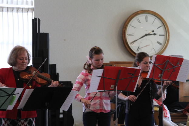Drei Frauen auf der Bühne mit Violinen spielend, mit Notenpulten und Noten davor, während ein Zuschauer im Vordergrund sitzt; eine Uhr ist an der weißen Wand im Hintergrund angebracht, und ein weißer Vorhang ist links sichtbar.
