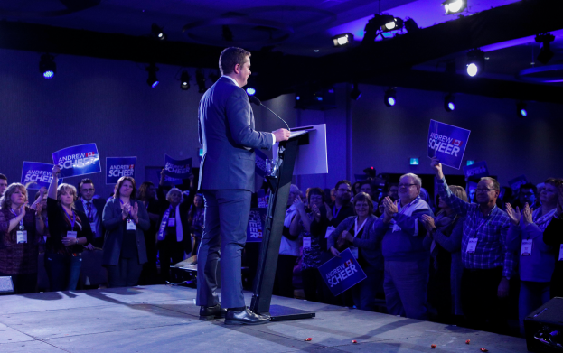 Ein Mann steht an einem Rednerpult mit Mikrofon und spricht zu einer Menge, wobei einige klatschen und Schilder halten, unter Deckenbeleuchtung auf einer Wahlveranstaltung.