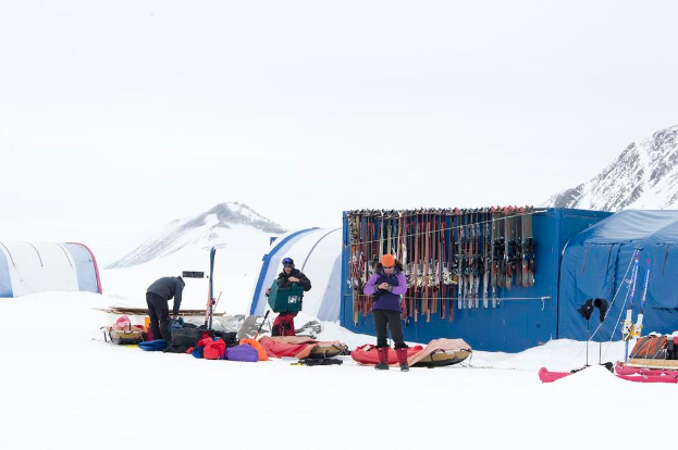 Drei Personen stehen auf einer verschneiten Landschaft mit Taschen drumherum, Zelte mit Skiern dahinter und schneebedeckten Hügeln im Hintergrund unter einem klaren Himmel.