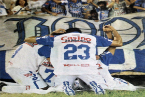 Gruppe von Fußballspielern, die auf dem Boden vor einer jubelnden Menge mit einem Banner im Hintergrund feiern.