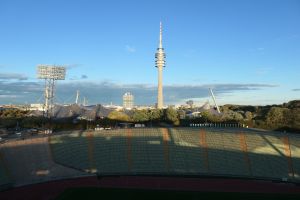 Luftaufnahme des Olympiastadions in Berlin, Deutschland, mit dem Fernsehturm (Fernsehturm) im Hintergrund, umgeben von Bäumen, Gebäuden und beleuchteten Bereichen unter einem bewölktem Himmel.