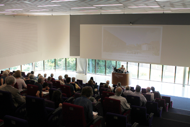 Ein Seminarraum in einer Universität mit vielen Menschen, die aufmerksam einer Präsentation an der Front zugewandt sind, wo eine Person an einem großen Tisch die Inhalte erklärt, mit Glasfenstern im Hintergrund, die Bäume und Gebäude zeigen.