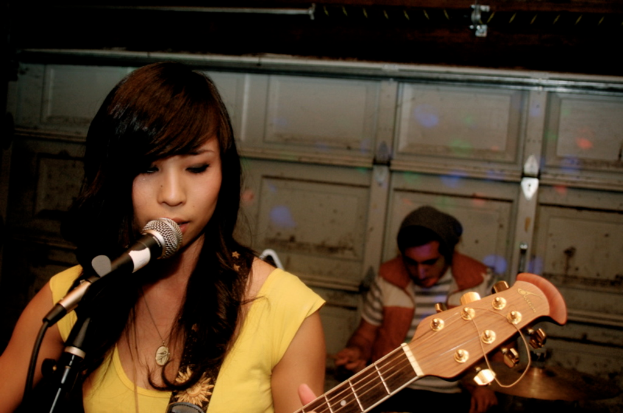 Eine Frau steht im Vordergrund und spielt Gitarre und singt in ein Mikrofon links, während ein Mann rechts im Hintergrund an einer Wand sitzt.