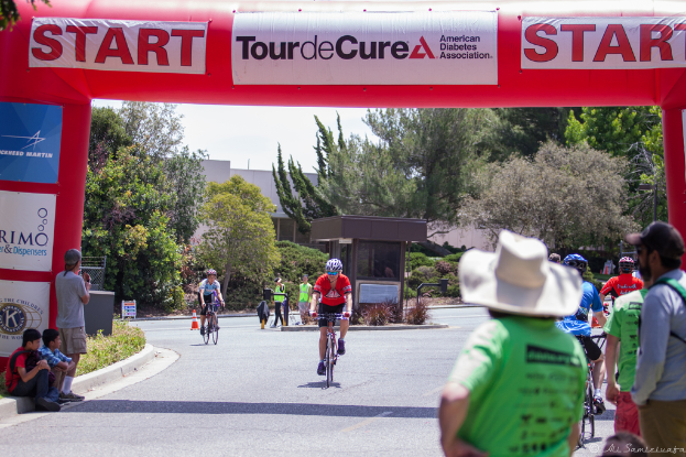 Eine belebte Straßenszene mit Menschen auf Fahrrädern, einige sitzen auf einem Gehweg und andere stehen auf der Straße, mit einer Hütte und umgeben von Pflanzen und Bäumen im Hintergrund.