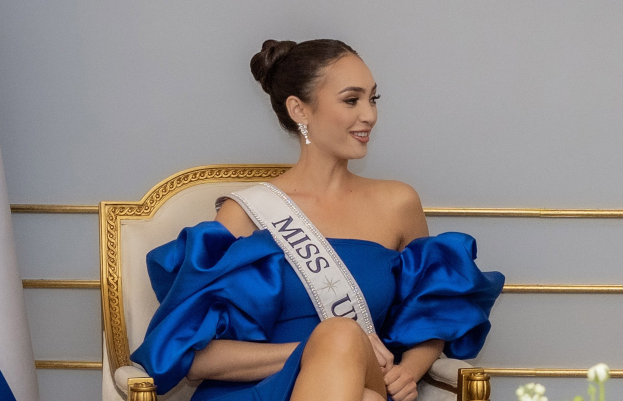 Frau in einem blauen Kleid mit einem "Miss Universe 2019"-Band, das auf einem Stuhl sitzt und von Blumen umgeben ist.