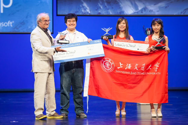 Vier Personen auf einer Bühne mit Auszeichnungen, Schecks und einer Fahne, die lächeln mit einem digitalen Bildschirm dahinter, der eine Projektion anzeigt.