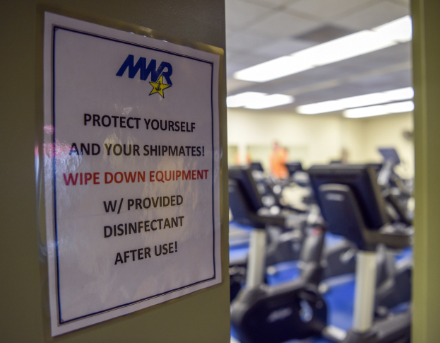 Ein Türschild mit der Aufschrift 'Schützen Sie sich und Ihre Schiffskameraden' mit Laufbändern und Deckenleuchten im Hintergrund, das Bild leicht unscharf, um Bewegung anzudeuten.